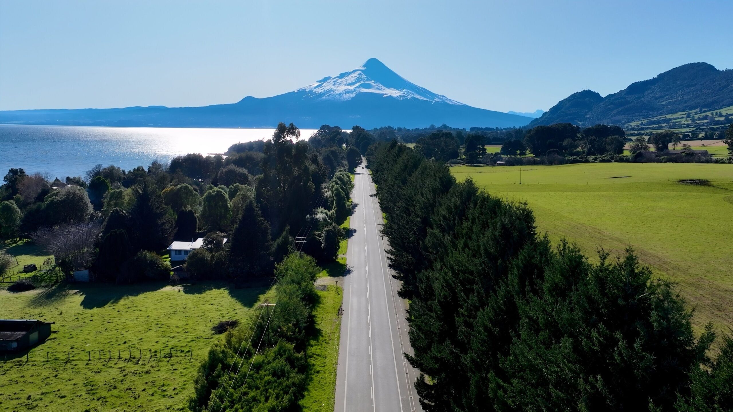 Camino bordeado de árboles junto al lago, con el volcán Osorno al fondo