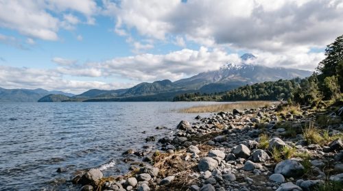 Orilla de piedras de un lago del sur de Chile con volcán nevado entre nubes al fondo
